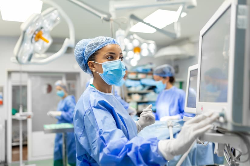 A nurse in blue scrubs and a surgical cap intently monitors data on a medical screen, operating the equipment with gloved hands. In the background, colleagues in similar attire are focused on their tasks in a bright operating room, filled with surgical lights and advanced equipment, reflecting a sterile and professional atmosphere.