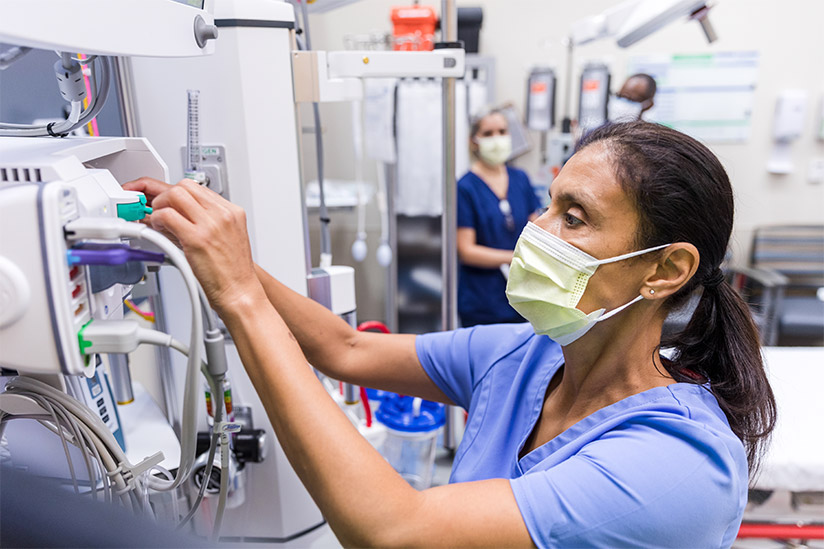 A female emergency room nurse wearing blue scrubs and a yellow surgical mask is checking monitoring equipment in preparation before receiving a trauma patient.