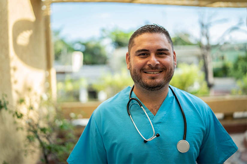 A confident male nurse is wearing blue scrubs and standing in an atrium area of a hospital. He is smiling and looking straight ahead.