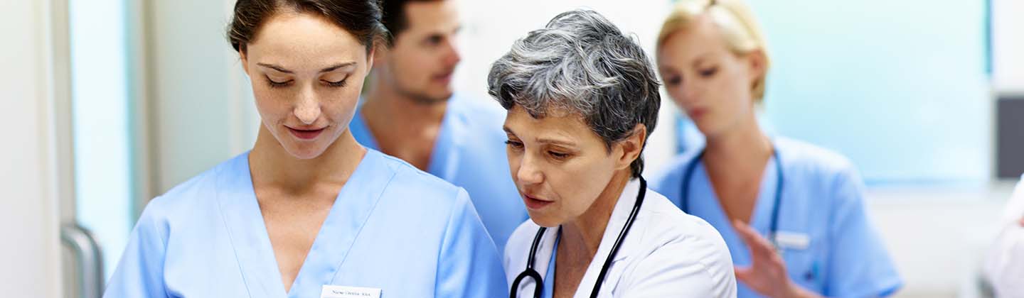 A group of nurses are in a hospital hallway walking and talking. An older nurse wearing a white lab coat is pointing to a clipboard a younger nurse is holding. She appears to be speaking about something that is documented on the clipboard while the younger nurse is listening. 