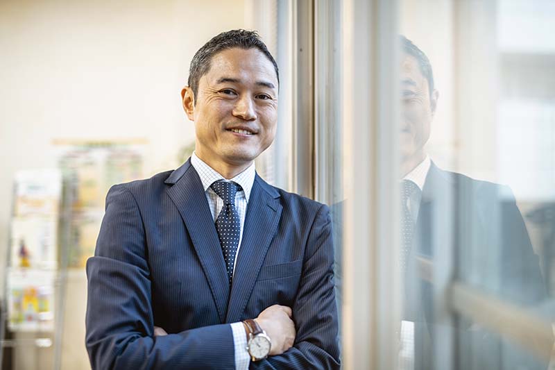 A smiling male nurse is wearing a dark blue suit. His arms are folded in front of him, and he is leaning against a glass windowpane.