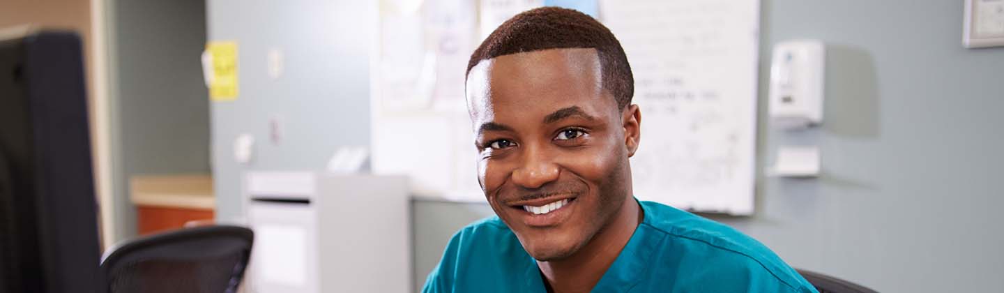 A smiling African American male nurse is seated at a computer workstation in a hospital ward. He is wearing blue scrubs and holding a clipboard and pen. 