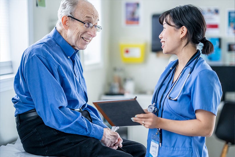 A nurse in a blue scrub top and badge provides coaching to a smiling elderly man in a blue shirt, holding a tablet in a clinic office, demonstrating a patient-centered approach in nursing.