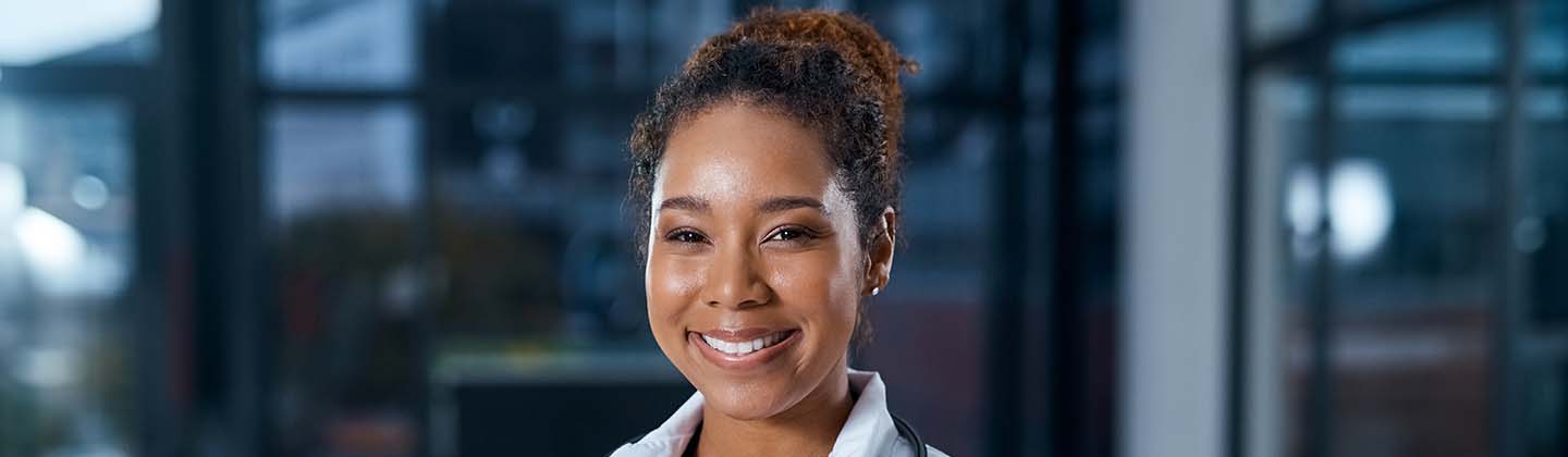 A confident African American nurse wearing scrubs and a while lab coat stands with her arms crossed in a darkened hospital hallway. She is smiling and looking straight ahead.