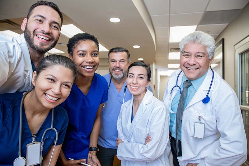 A group of healthcare workers are standing in a hallway and are gathered for a picture. They are leaning into the camera and smiling.