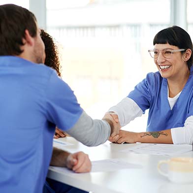 A%20group%20of%20nurses%20wearing%20blue%20scrubs%20are%20seated%20around%20a%20conference%20table%20and%20smiling.%20Two%20nurses%20are%20shaking%20hands%20across%20the%20table.