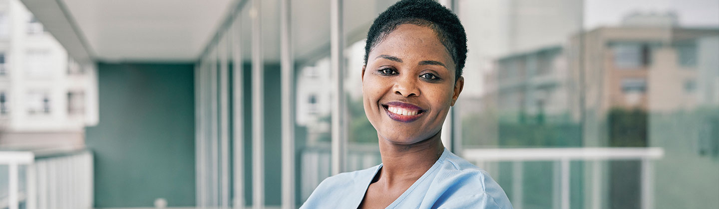 A smiling female nurse is standing in front of her hospital with her arms crossed. 