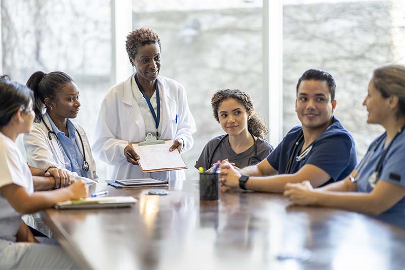 A female nursing leader is standing at a conference table, engaged in conversation with a group of her staff, who are seated around the table.