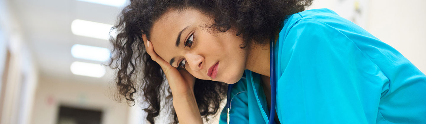 A female nurse wearing light blue scrubs is seated alone in a hospital hallway and holding her head in her hands. She looks concerned and tired.