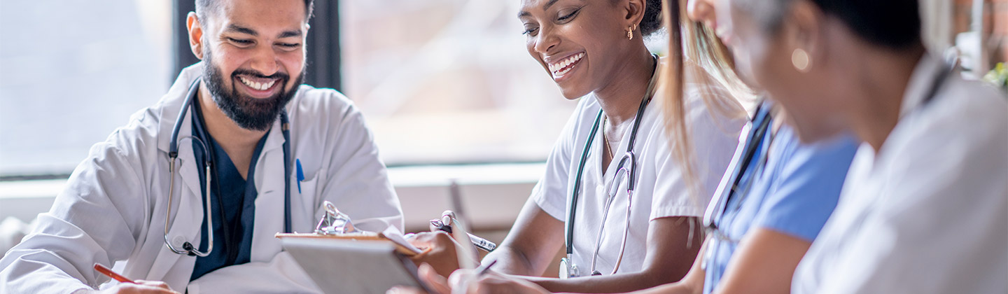 A small group of four medical professionals sit around a boardroom table as they meet to discuss patient cases. They are each dressed professionally in scrubs and lab coats as they focus on working together.