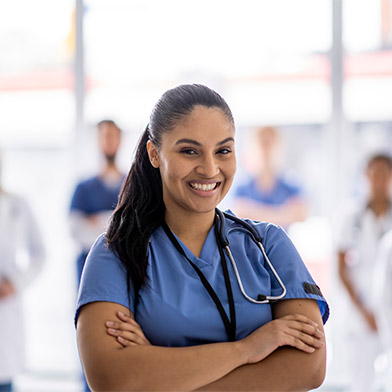 A%20female%20bi-racial%20nurse%20stands%20with%20her%20arms%20crossed%20in%20front%20of%20her%20and%20a%20smile%20on%20her%20face,%20as%20she%20poses%20for%20a%20portrait.%20She%20is%20wearing%20scrubs,%20and%20her%20team%20can%20be%20seen%20standing%20with%20her%20in%20the%20background.